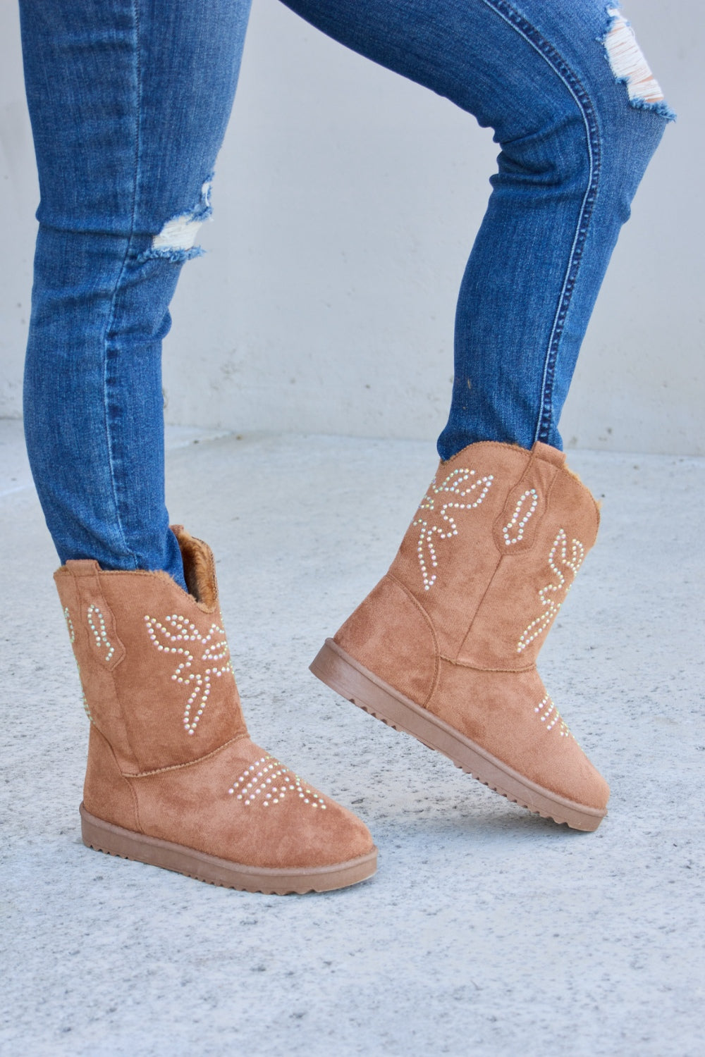 A person wearing ripped blue jeans and Forever Link Rhinestone Furry Flat Boots stands on a concrete surface.