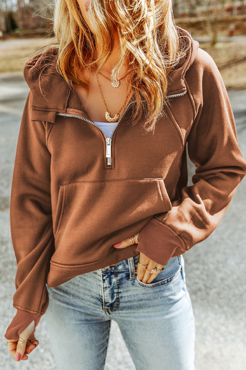 A person with long hair is walking away wearing a Brown Quarter Zip Kangaroo Pocket Hoodie and light blue ripped jeans. The background shows an outdoor setting.