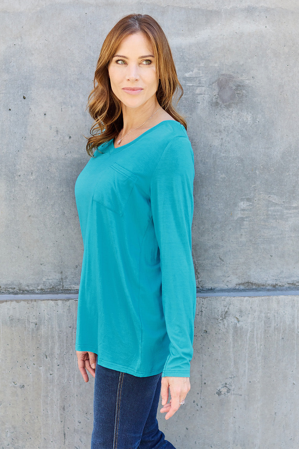 A woman with light brown hair stands against a concrete wall, wearing the Basic Bae Full Size V-Neck Long Sleeve Top in olive green and blue jeans, with a white backpack on her shoulder. Her outfit exudes basic style and is made from polyester, ensuring it’s both comfortable and suitable for machine wash cold. She is looking at the camera.