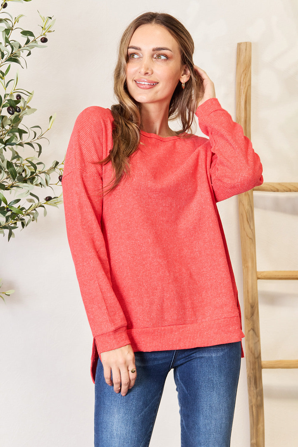 A woman wearing the Basic Bae Round Neck Drop Shoulder Slit Sweatshirt in red and blue jeans stands indoors, smiling and touching her hair, with a decorative plant and wooden ladder in the background.