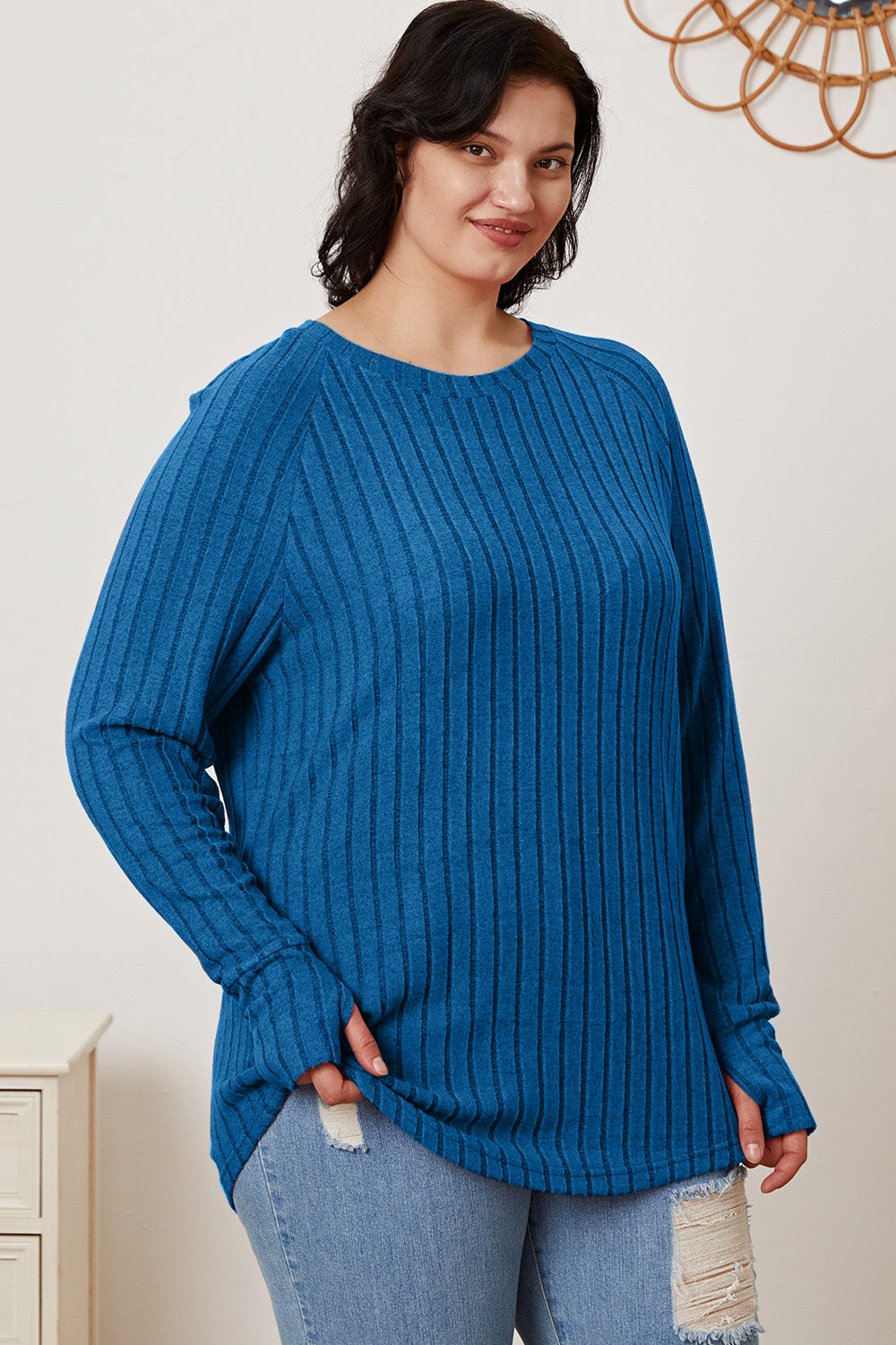 A person wearing the Basic Bae Full Size Ribbed Thumbhole Sleeve T-Shirt in blue and light-washed jeans stands smiling, with a decorative wall hanging in the background.