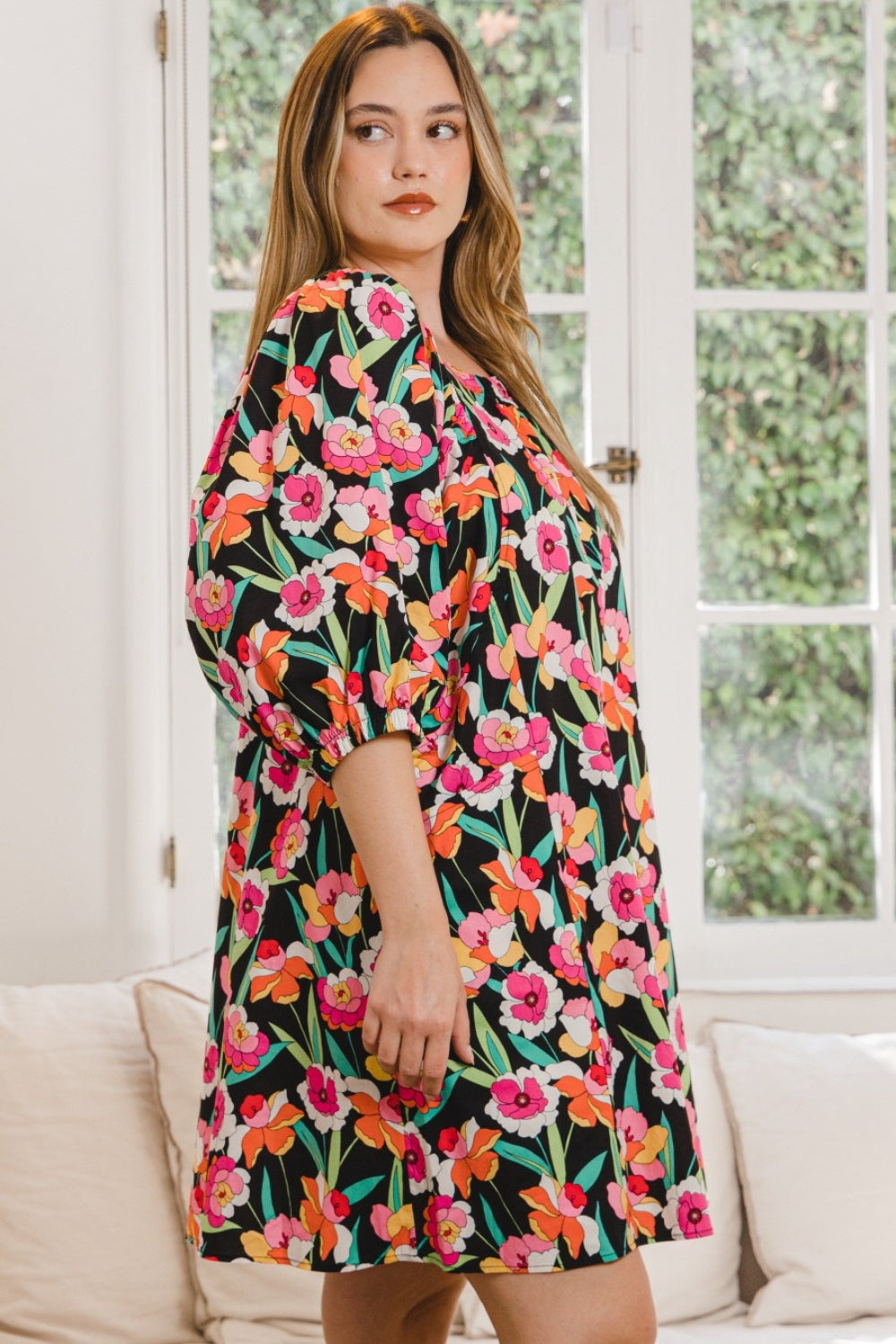 A woman wearing the stylish ODDI Full Size Floral Puff Sleeve Mini Dress stands indoors, framed by a large window with lush greenery outside.