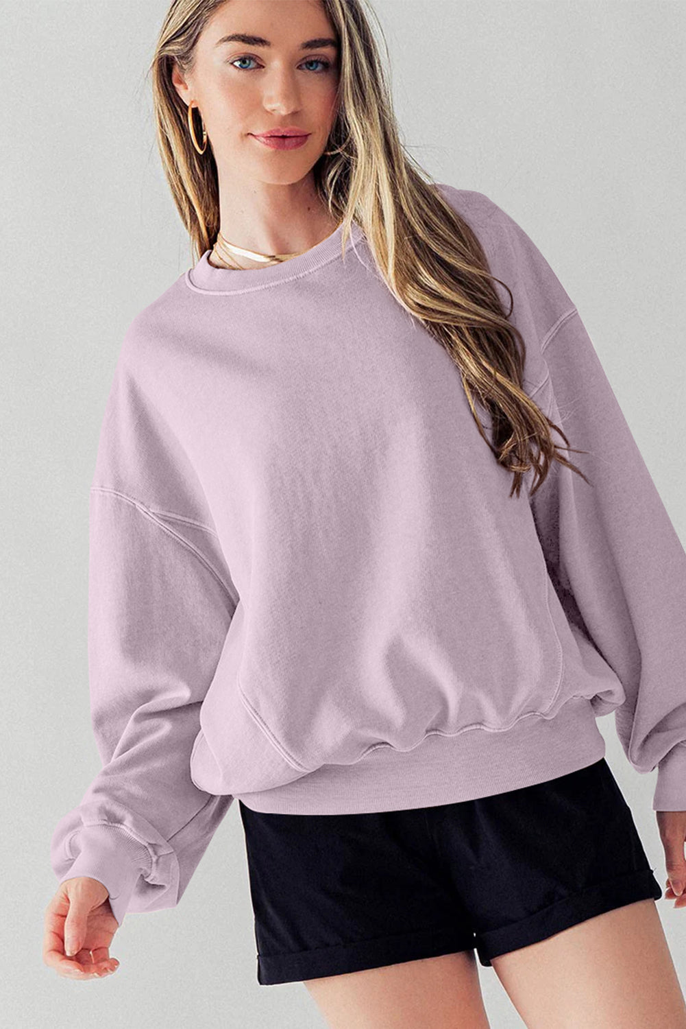A woman facing away wears the Orchid Petal Exposed Seam Batwing Sleeve Drop Shoulder Sweatshirt and black shorts. She poses with her right arm raised, resting on her head.