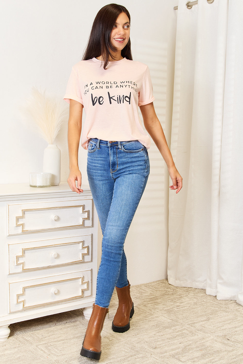 A person wears a Simply Love Slogan Graphic Cuffed T-Shirt in light pink, adorned with the inspiring message, "In a world where you can be anything, be kind," and featuring stylish cuffed sleeves.