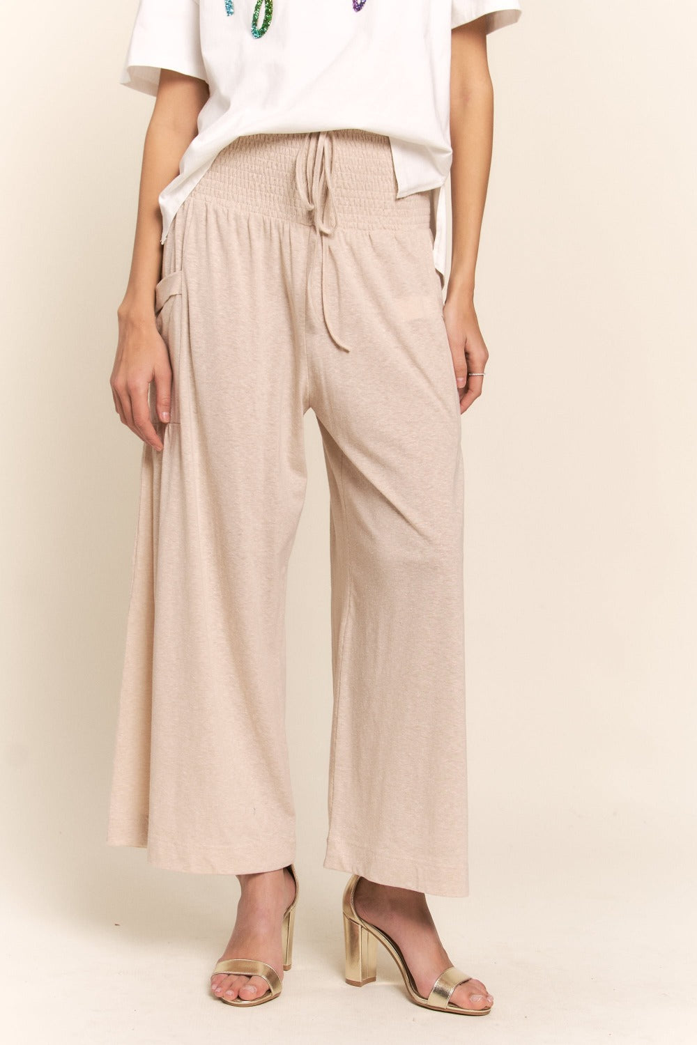 A person wearing J.NNA Smocked Waist Boho Wide Leg Pants with Pockets in beige stands in low-heeled sandals against a plain background, paired with a chic white T-shirt.