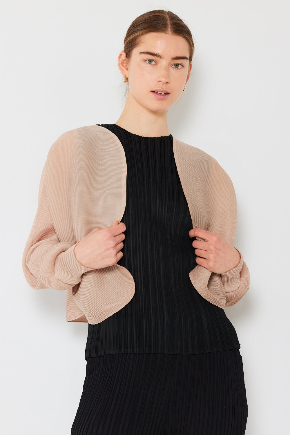 A person stands wearing the stylish Marina West Swim Rib Pleated Puff Sleeve Bolero Cardigan in beige, featuring draped sleeves, paired with a trendy black pleated top.