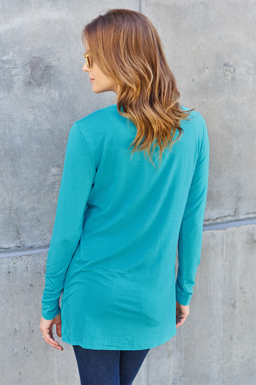 A woman with long hair wearing sunglasses, a Basic Bae Full Size Round Neck Long Sleeve Top in turquoise, and jeans is standing in front of a concrete wall with one hand on her hip.