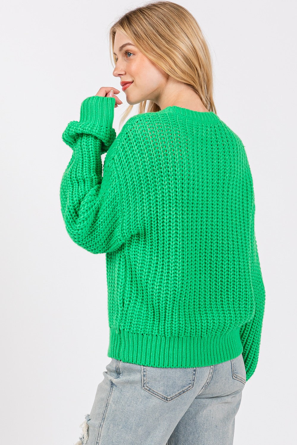 A person wearing a SAGE + FIG round neck drop shoulder sweater, ripped light blue jeans, and a green hat poses against a white background.