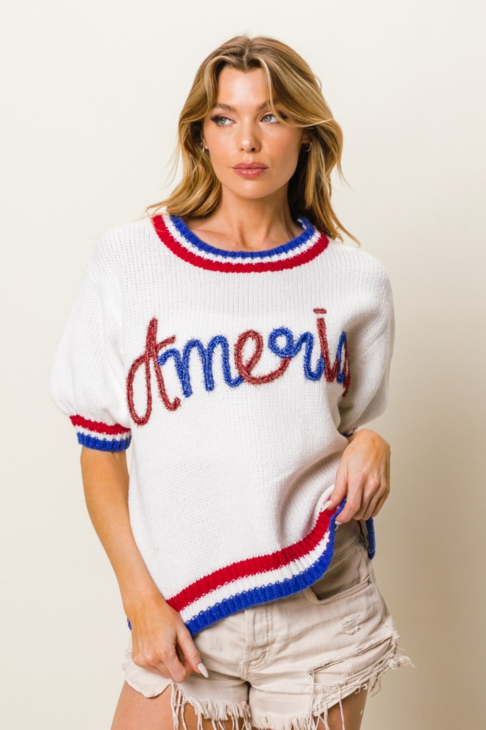 A person wearing the BiBi Metallic America Letter Short Sleeve Sweater, featuring "Amour" written in red, blue, and brown letters, paired with white shorts. The sweater is accented by red and blue trim on the sleeves and neckline.