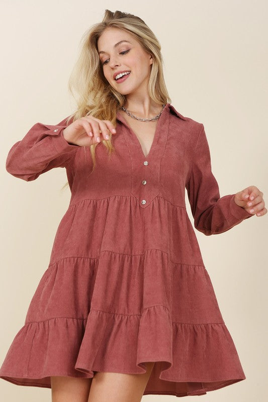 The woman in the Corduroy tiered dress, featuring a deep V-neck, smiles as she gently touches her forehead, showcasing the light blue fabric cascading elegantly.