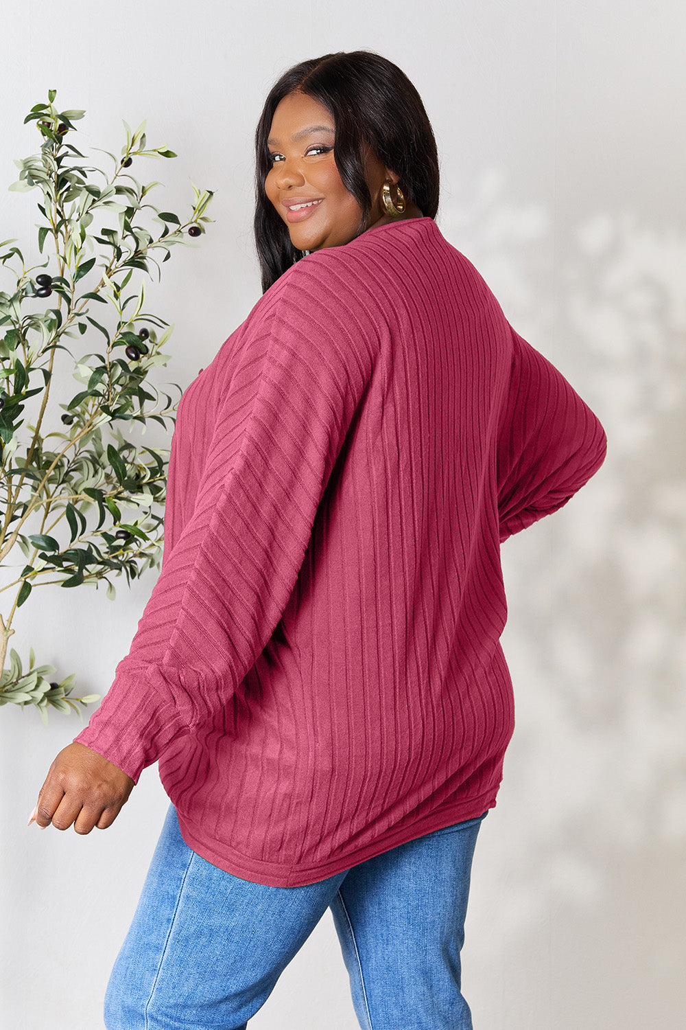 A person with wavy hair, wearing the Basic Bae Full Size Ribbed Cocoon Cardigan over a white top and beige pants, stands in front of a light-colored background with a leafy plant, showcasing an effortlessly basic style.