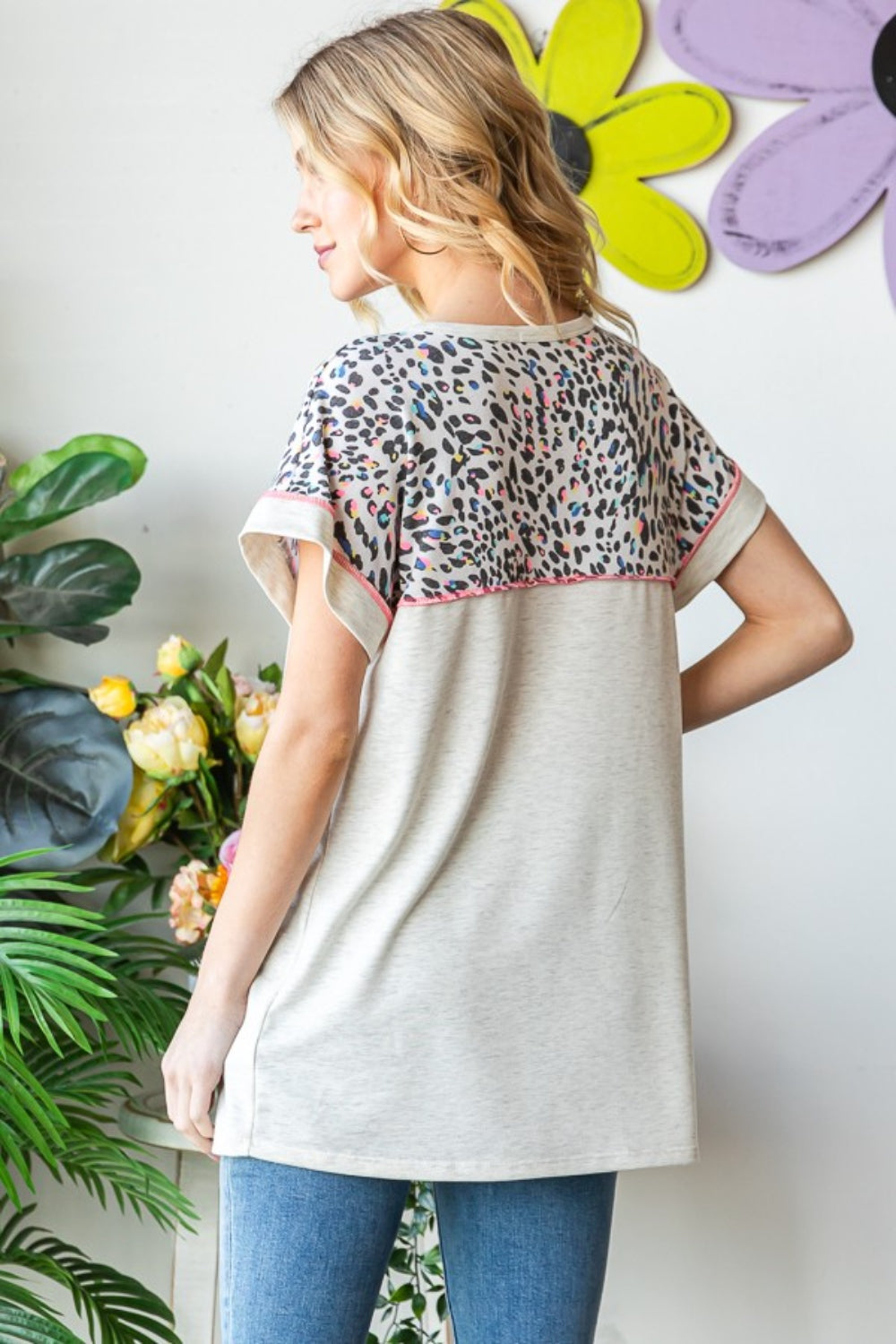 A woman stands indoors wearing a Heimish Full Size Exposed Seam Leopard Short Sleeve T-Shirt with blue jeans, holding the hem of her shirt. She is surrounded by green plants and a wall adorned with yellow and purple flower decorations.