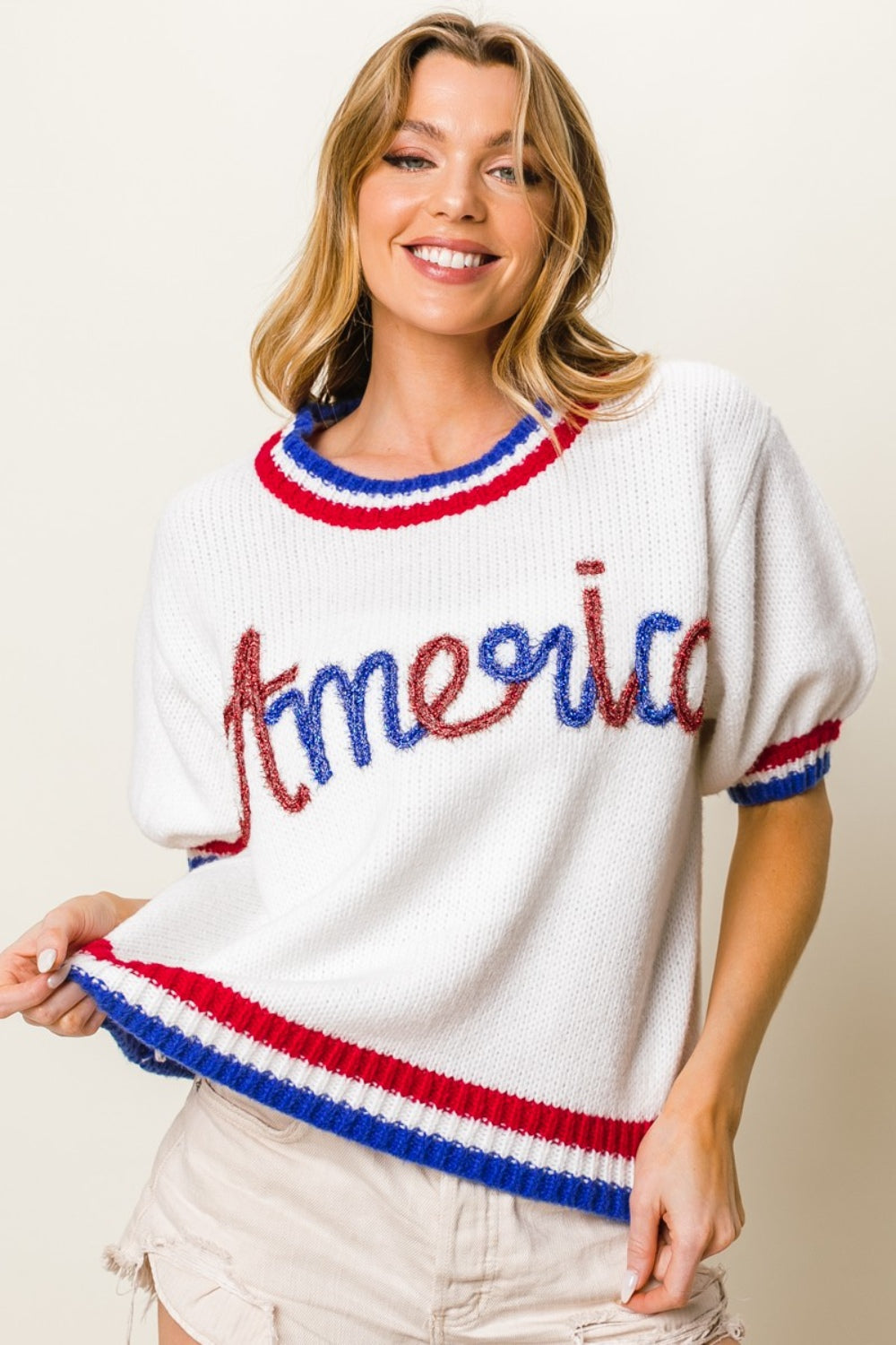 A person wearing the BiBi Metallic America Letter Short Sleeve Sweater, featuring "Amour" written in red, blue, and brown letters, paired with white shorts. The sweater is accented by red and blue trim on the sleeves and neckline.