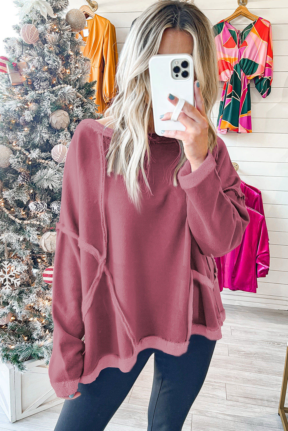 A woman with long hair is wearing an Exposed Seam Drop Shoulder Raw Hem Oversized Sweatshirt in pink, paired with denim shorts, standing indoors in front of a potted plant and a decorative wall.