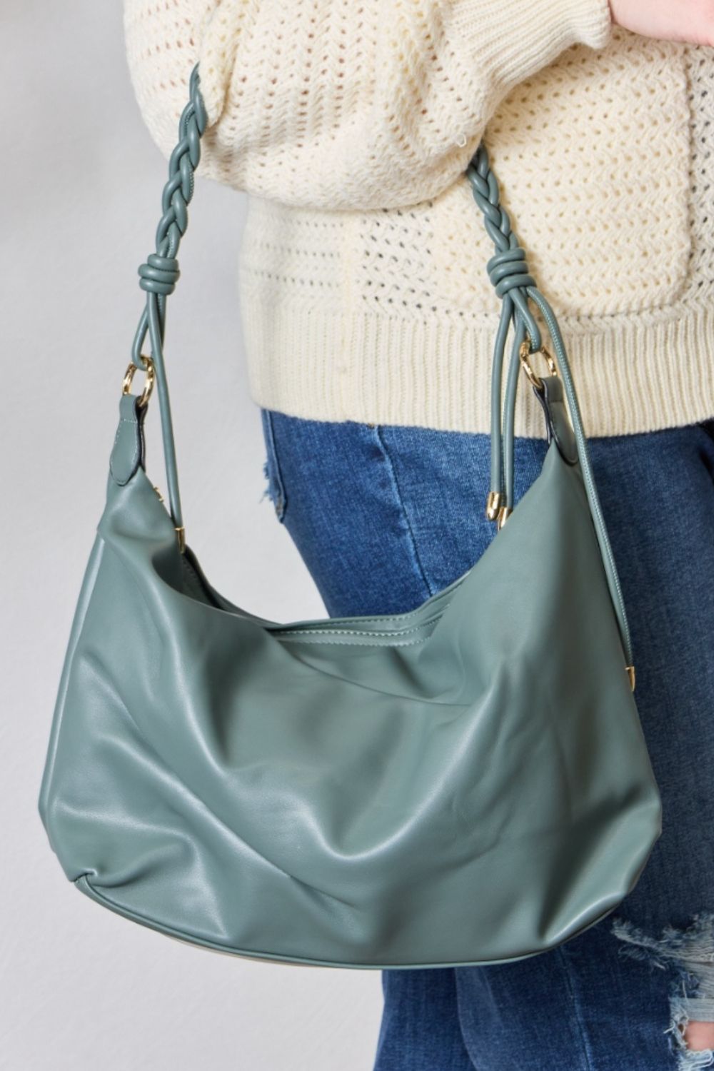 A person in a knitted sweater and jeans holds the SHOMICO Braided Strap Shoulder Bag, a blue-green statement accessory featuring straps crafted from vegan leather.