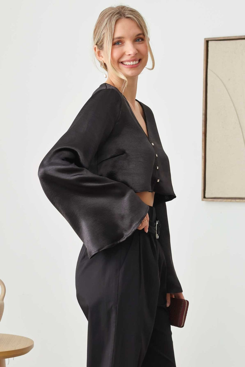 A person in a black long-sleeve Mustard Seed Satin Crop Top with rhinestone button closures and pants, holding a clutch, stands near a wooden table with a vase. Two framed artworks elegantly hang on the wall behind.