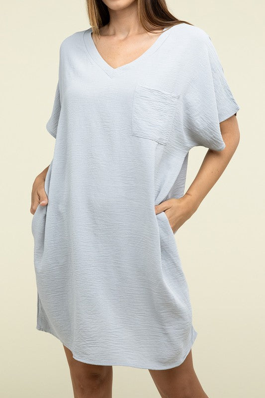 A woman wearing a loose light brown Woven Airflow V Neck T-Shirt Dress with Pockets stands against a plain background. Made from breathable fabric, this summer dress includes pockets where her hands rest as she smiles slightly.