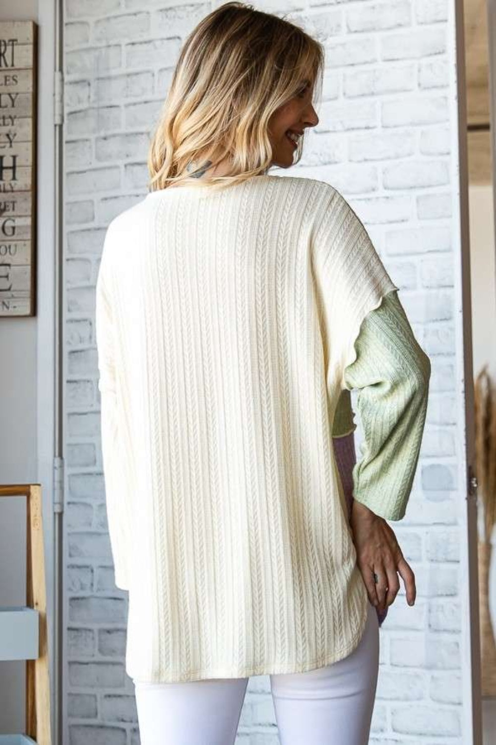 A person dressed in the Culture Code Color Block Round Neck Dropped Shoulder Blouse, featuring pastel shades of green, ivory, and lavender, stands in a room with a decorative sign on the brick wall.