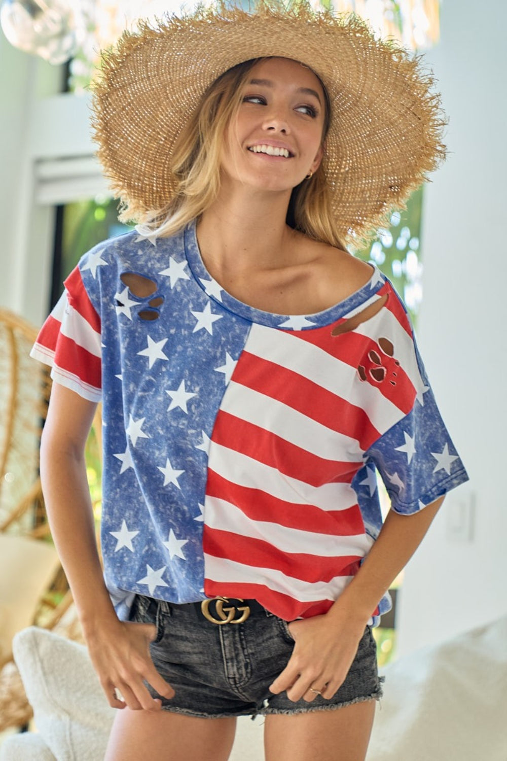 A person wearing a large straw hat stands indoors, smiling. They are also proudly sporting the BiBi American Flag Theme Short Sleeve T-Shirt with denim shorts and a black belt with a gold buckle.