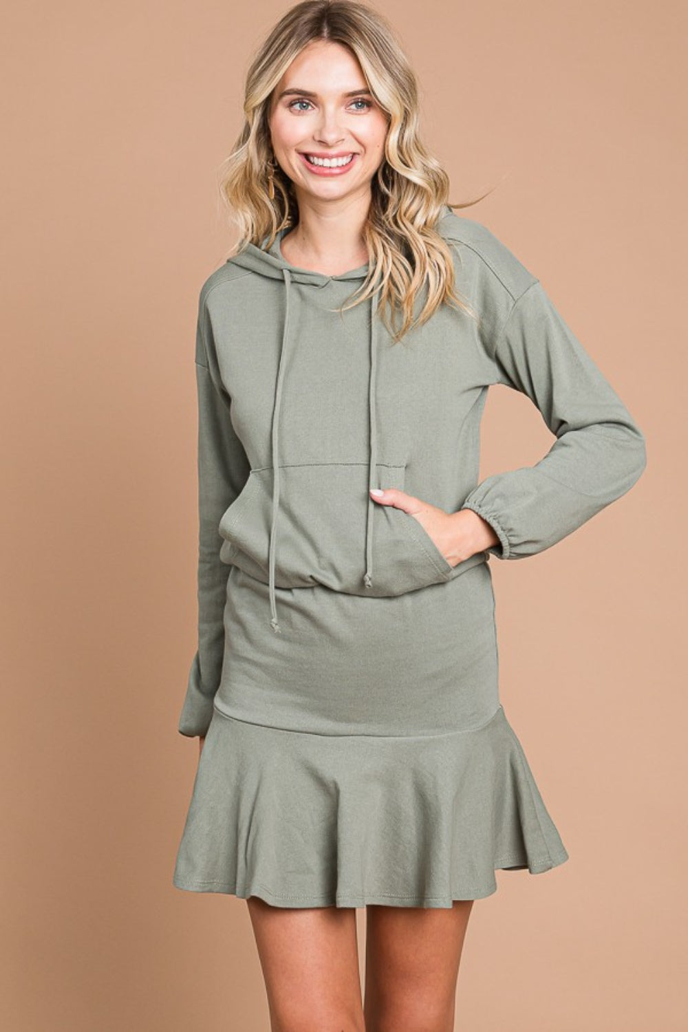 A woman stands smiling against a beige background, wearing the Culture Code Full Size Drawstring Dropped Shoulder Hooded Mini Dress.