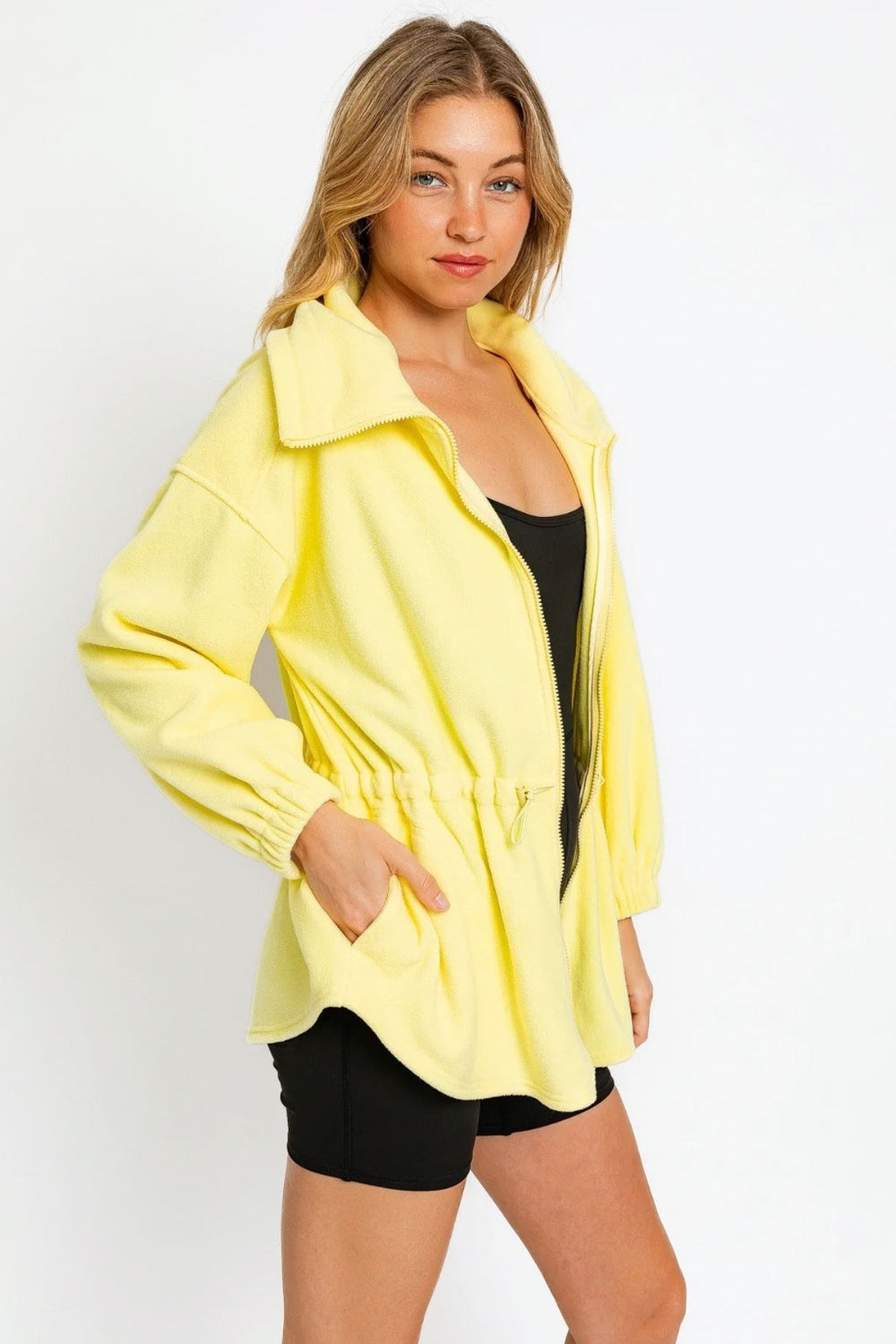 A woman wearing a black outfit and a yellow Le Lis Zip Up Waist Drawstring Soft Fleece Jacket stands against a plain white background.