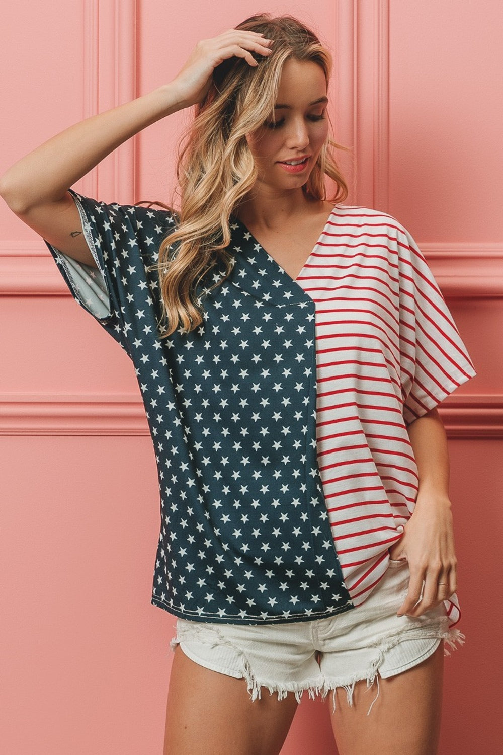 A woman stands against a pink wall, wearing the BiBi US Flag Themed Color Block Short Sleeve T-Shirt, which features one half blue with white stars and the other half red and white stripes. She is smiling with long wavy hair, showcasing her American pride casual wear.