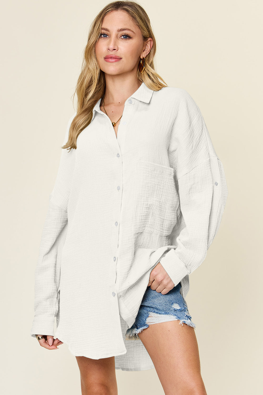 A person is wearing the Double Take Full Size Pocketed Texture Button Up Shirt, a long-sleeved, color-blocked piece made of 100% cotton in shades of green and cream. They have paired it with distressed denim shorts and are standing against a neutral backdrop. The shirt features buttoned cuffs and offers a relaxed yet stylish look. It is easy to care for with machine wash cold instructions.