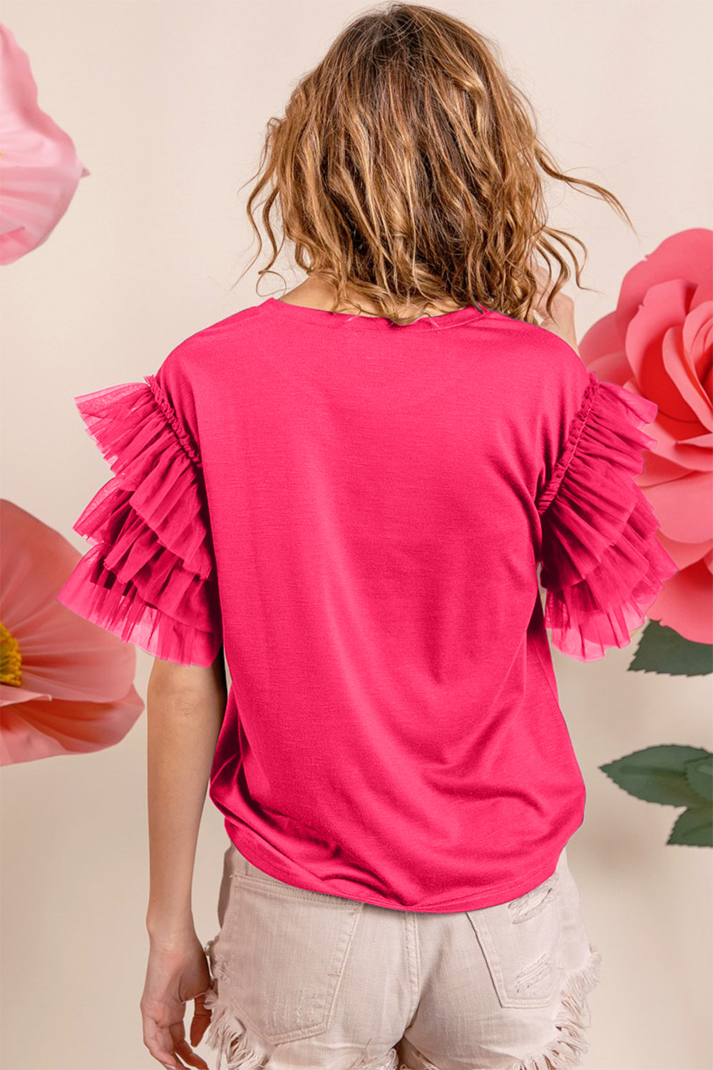 A smiling woman with long hair sits on a bed, wearing a BiBi Ruffle Mesh Lace Layered Short Sleeve Top in bright pink.