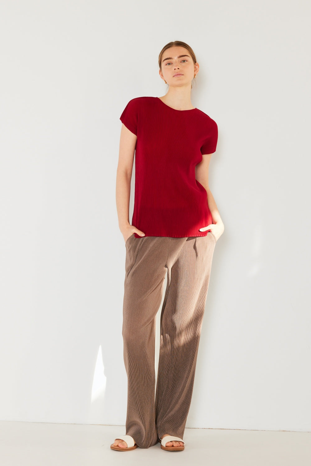 A person stands against a plain background, wearing a Marina West Swim Rib Pleated Cap Sleeve Top in red and brown pants, with hands in pockets.