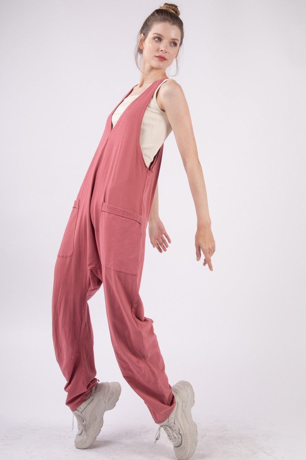 Someone is standing against a plain backdrop, wearing the VERY J Plunge Sleeveless Jumpsuit in pink and white shoes, with their hands casually tucked into the jumpsuit's pockets.