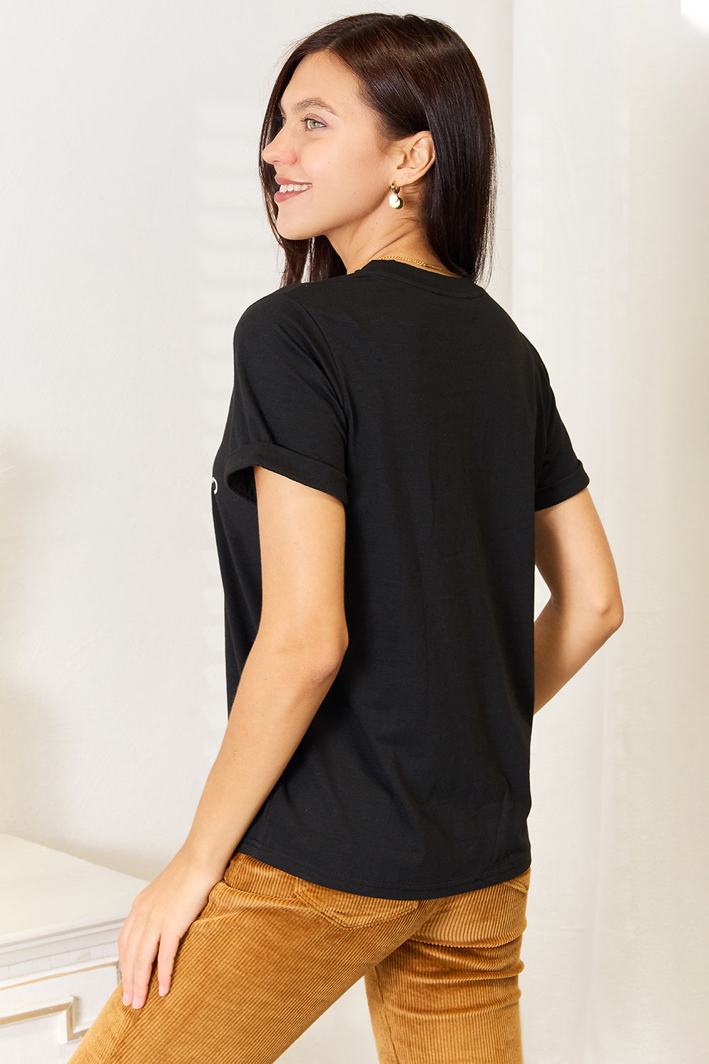 A woman with long dark hair smiles while wearing a trendy black Simply Love Letter Graphic Round Neck T-Shirt featuring the text "ME? Sarcastic? NEVER" and brown corduroy pants. She stands in a room with white walls and decor.