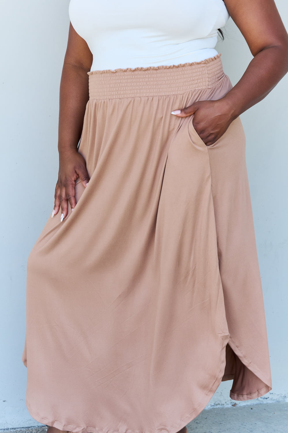 A person stands against a wall, wearing a white sleeveless top, the Doublju Comfort Princess Full Size High Waist Scoop Hem Maxi Skirt in Tan, and blue platform sandals. The right hand rests on the side of the skirt.