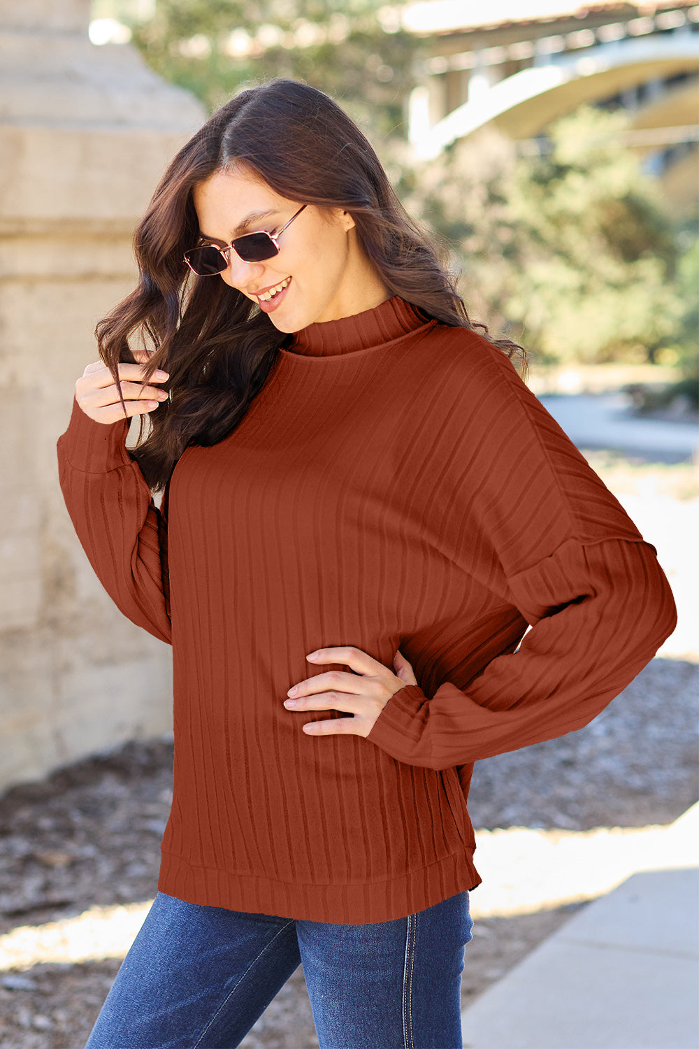 A woman with long hair is wearing a green Basic Bae Full Size Ribbed Exposed Seam Mock Neck Knit Top, blue jeans, and sunglasses. She is standing outdoors and smiling.