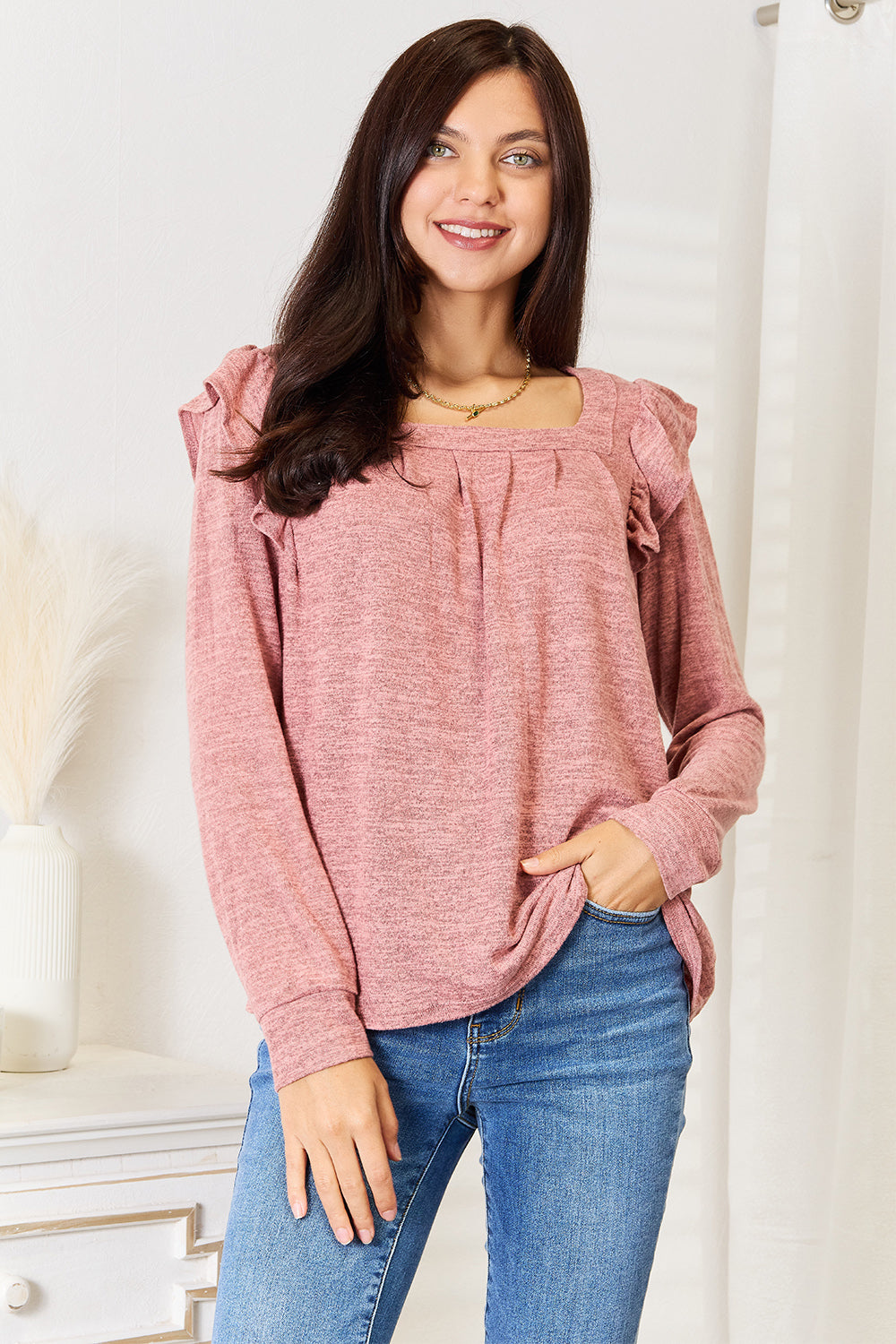 A woman wearing a Double Take Square Neck Ruffle Shoulder Long Sleeve T-Shirt in pink and blue jeans stands with one hand in her pocket, smiling. She has long curly hair and wears a necklace and bracelet. She stands in a room with white decor.