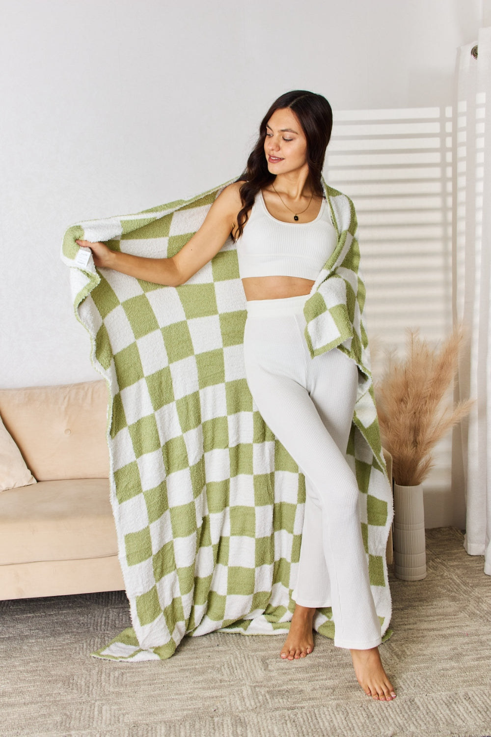 A person stands holding a large Cuddley Checkered Decorative Throw Blanket, smiling, in a room with a sofa and a potted plant. The blanket features a black and white checkerboard pattern, is made from 100% polyester, and imported for quality.