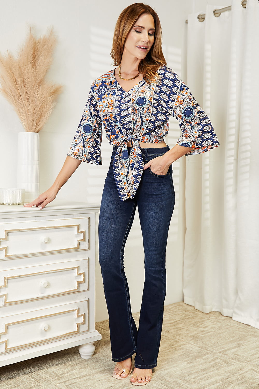 A woman dressed in a Tie Hem V-Neck Three-Quarter Sleeve Blouse and black pants stands indoors beside a white piece of furniture decorated with pampas grass.