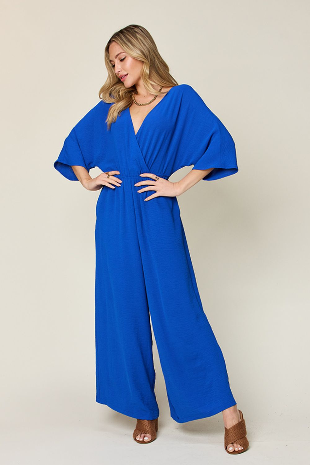 Person wearing the Double Take Full Size Surplice Wide Leg Jumpsuit with Pockets in royal blue, featuring wide legs, a deep V-neckline, and ruched detailing, posing with hands on hips against a plain light-colored background.