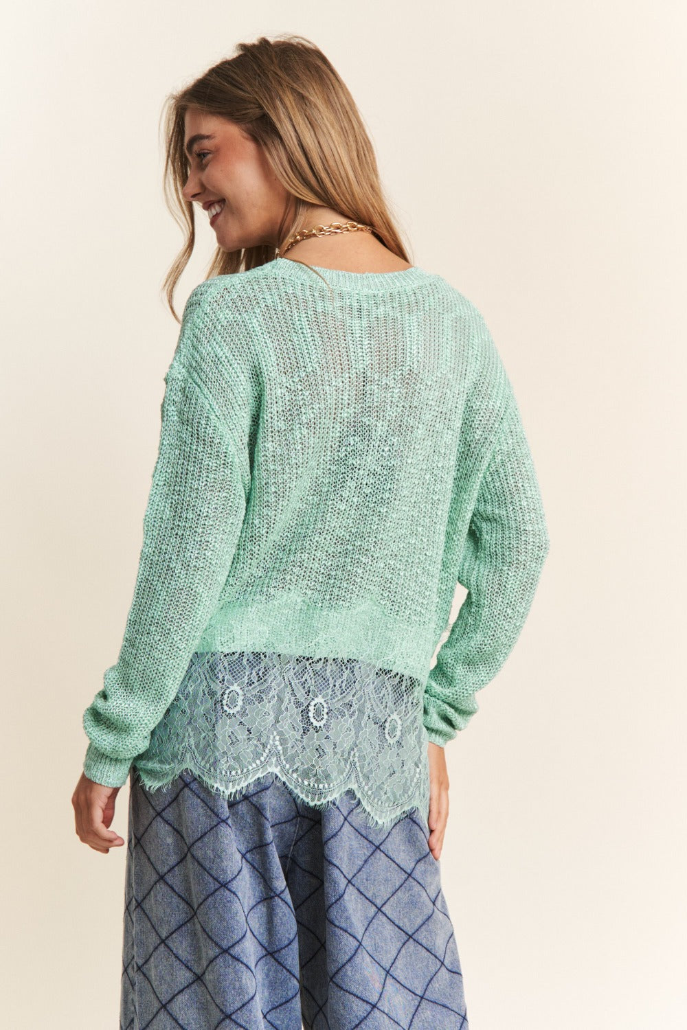 A woman with long hair is wearing the J.NNA Lace Detail Long Sleeve Knit Top in a versatile light green, paired with a lace trim shirt underneath and patterned blue pants. She has a neutral expression and her right hand near her face.