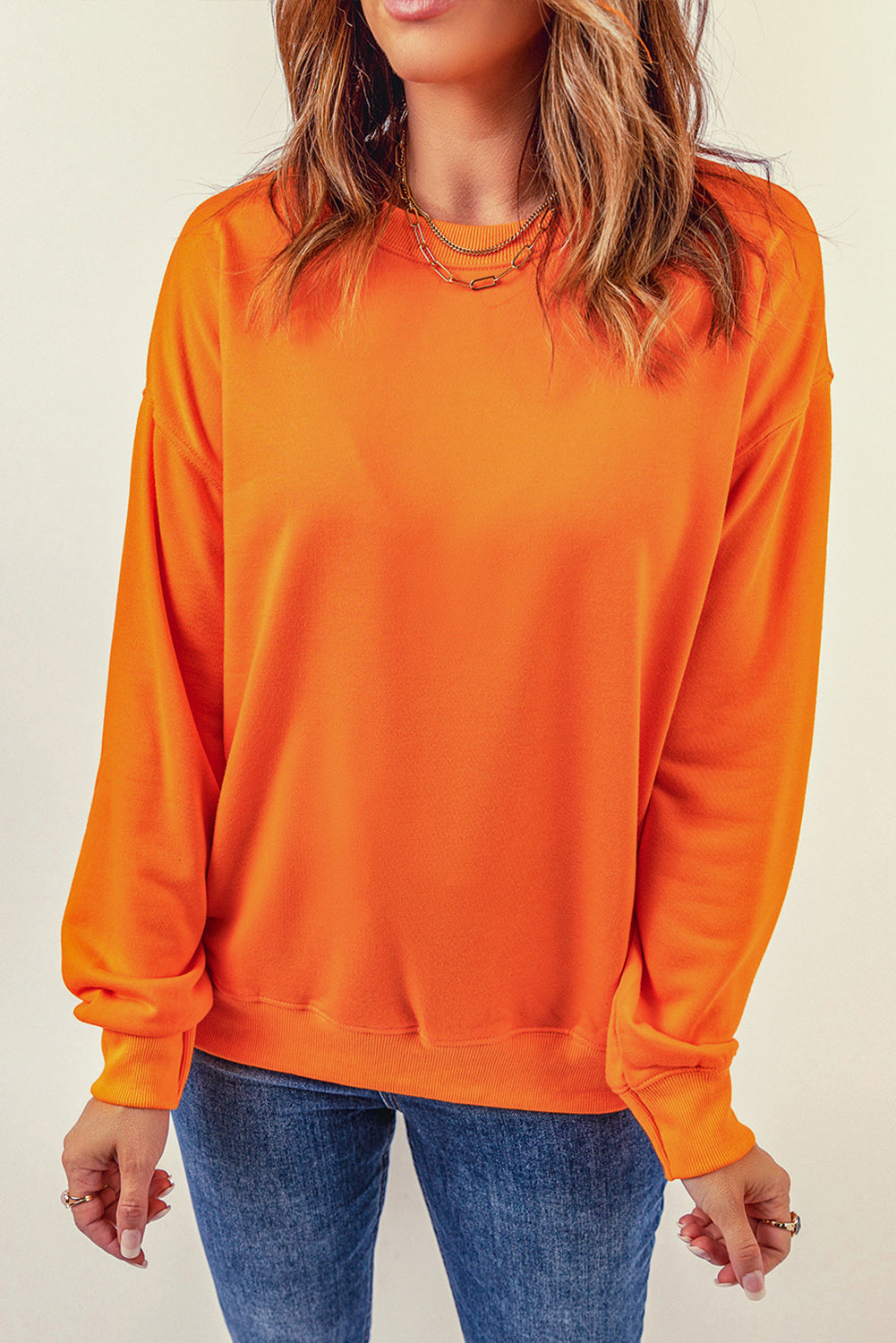 Person with wavy, shoulder-length hair wearing an Orange Plain Crew Neck Pullover Sweatshirt and blue jeans, standing with hand on hip, facing away.