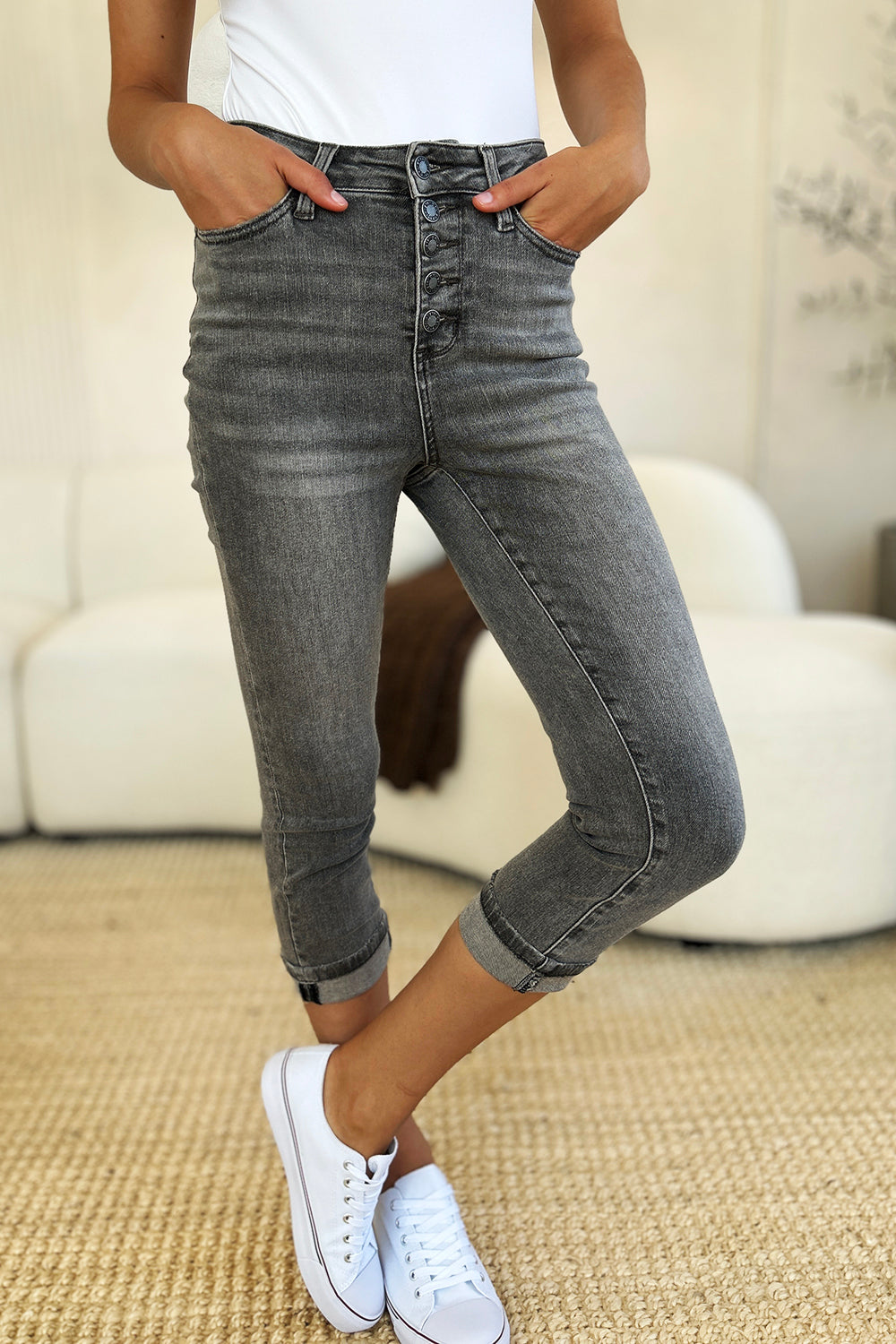 A person wearing Judy Blue Full Size Button Fly High Waist Cuffed Capris in gray, paired with a white top and white sneakers, stands on a beige carpeted floor.