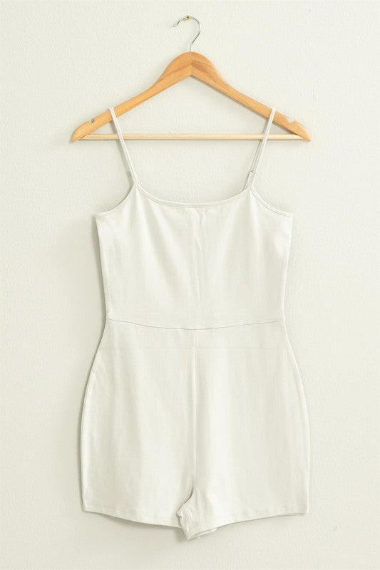 The Loving Me Fitted Sleeveless Romper, featuring a fitted bodice and spaghetti straps in black, hangs gracefully on a wooden hanger against a white wall.