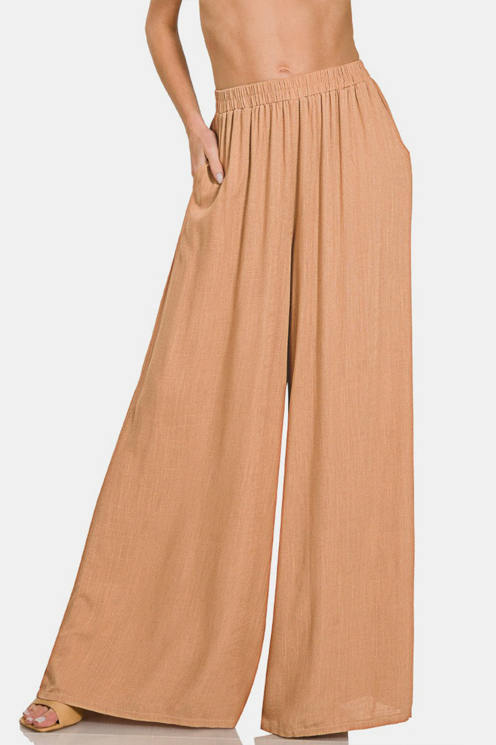 A person wearing Zenana Pleated Linen Blend Wide Leg Pants in tan, with hands in pockets, standing against a plain white background. Upper body and face are not visible.