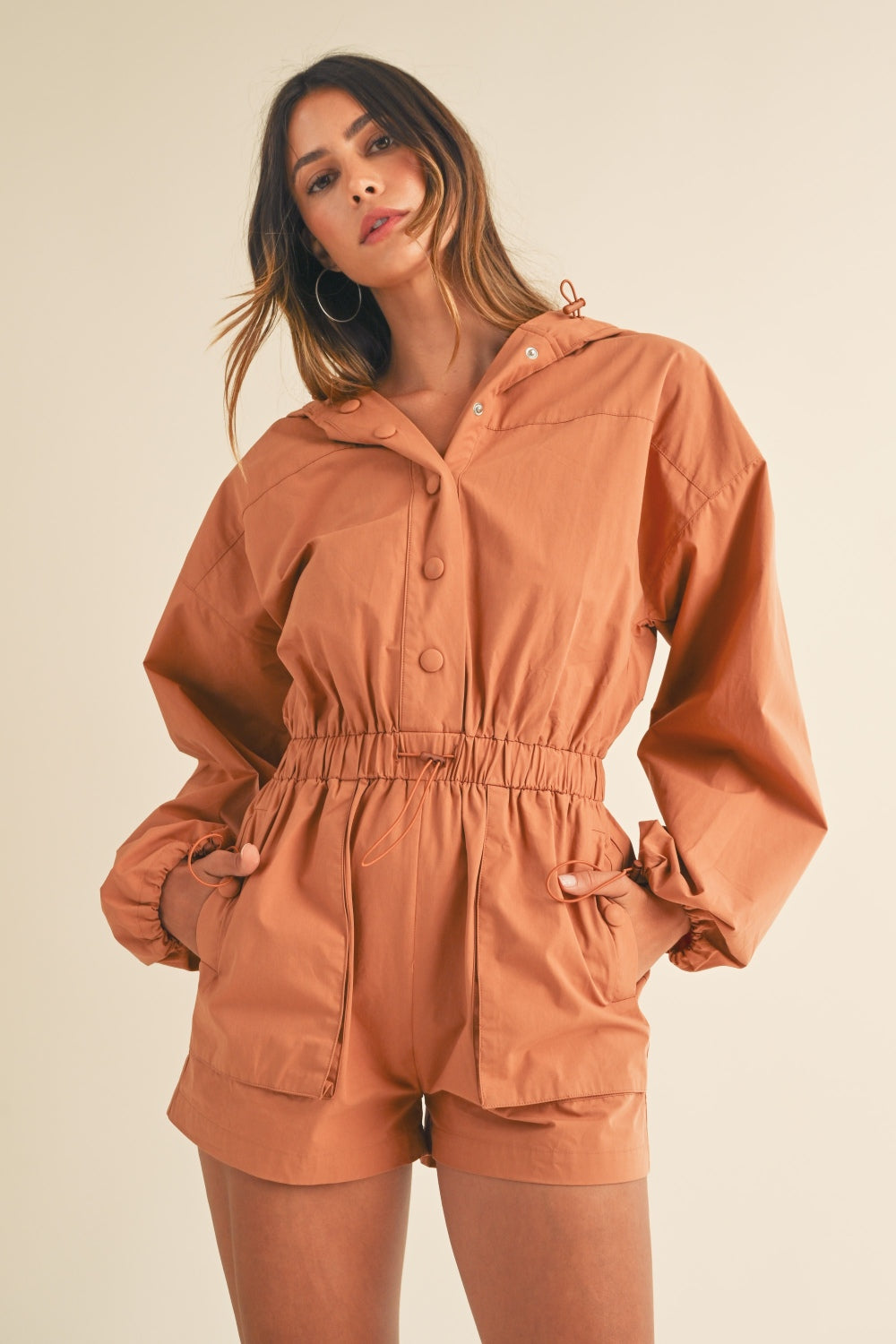 A person wearing the Mable Snap Down Long Sleeve Hooded Romper, featuring a brown color, long sleeves, a snap-down front, and an elastic waistband, is standing against a plain background.