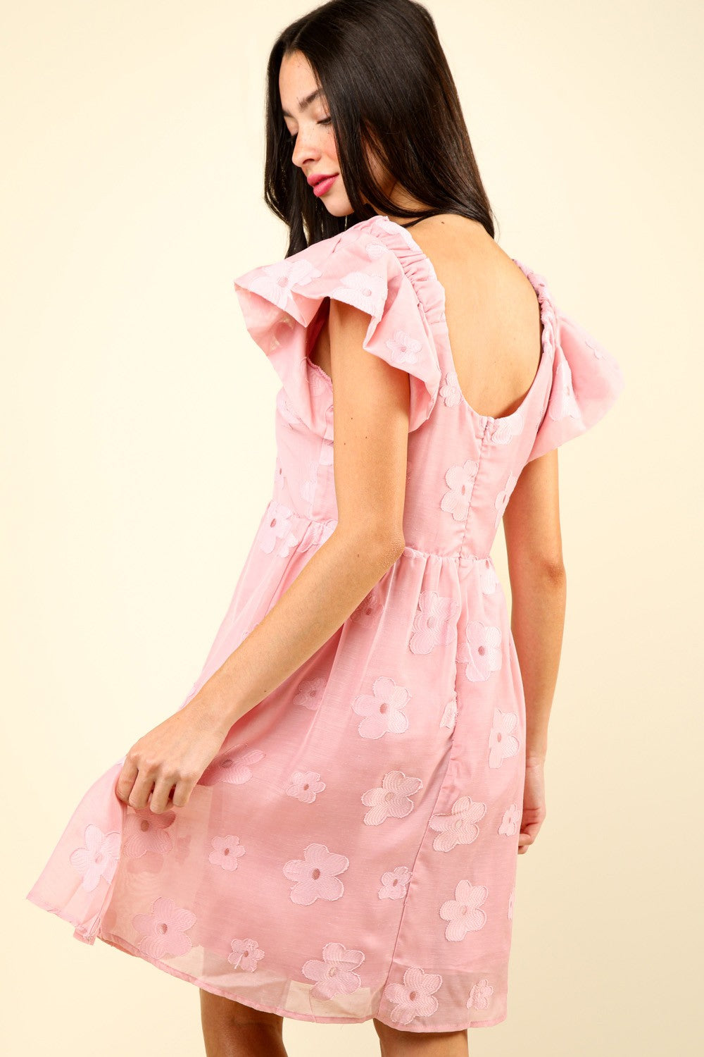 Woman in a VERY J Flower Embroidered Organza Mini Dress with flutter sleeves stands against a light background, resting one hand on the skirt and the other slightly bent. She is wearing strappy sandals.