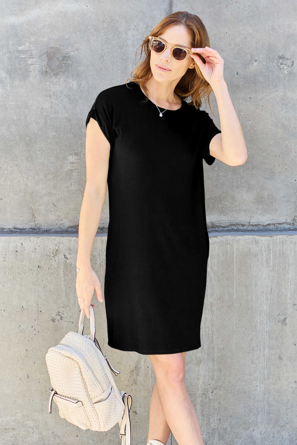 A woman wearing a Basic Bae Bamboo Full Size Round Neck Short Sleeve Dress with Pockets and sunglasses holds the strap of a light-colored backpack while standing against a concrete wall.