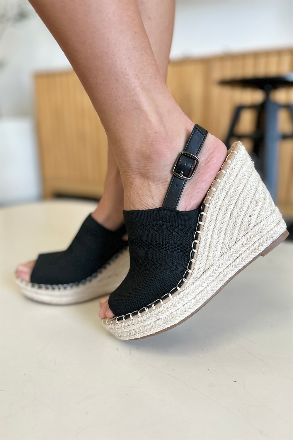 Close-up of a person's feet wearing chic black Forever Link Peep Toe High Heel Sandals with a braided espadrille base and adjustable heel straps. The background is blurred, making these sandals the stylish choice for any outfit.