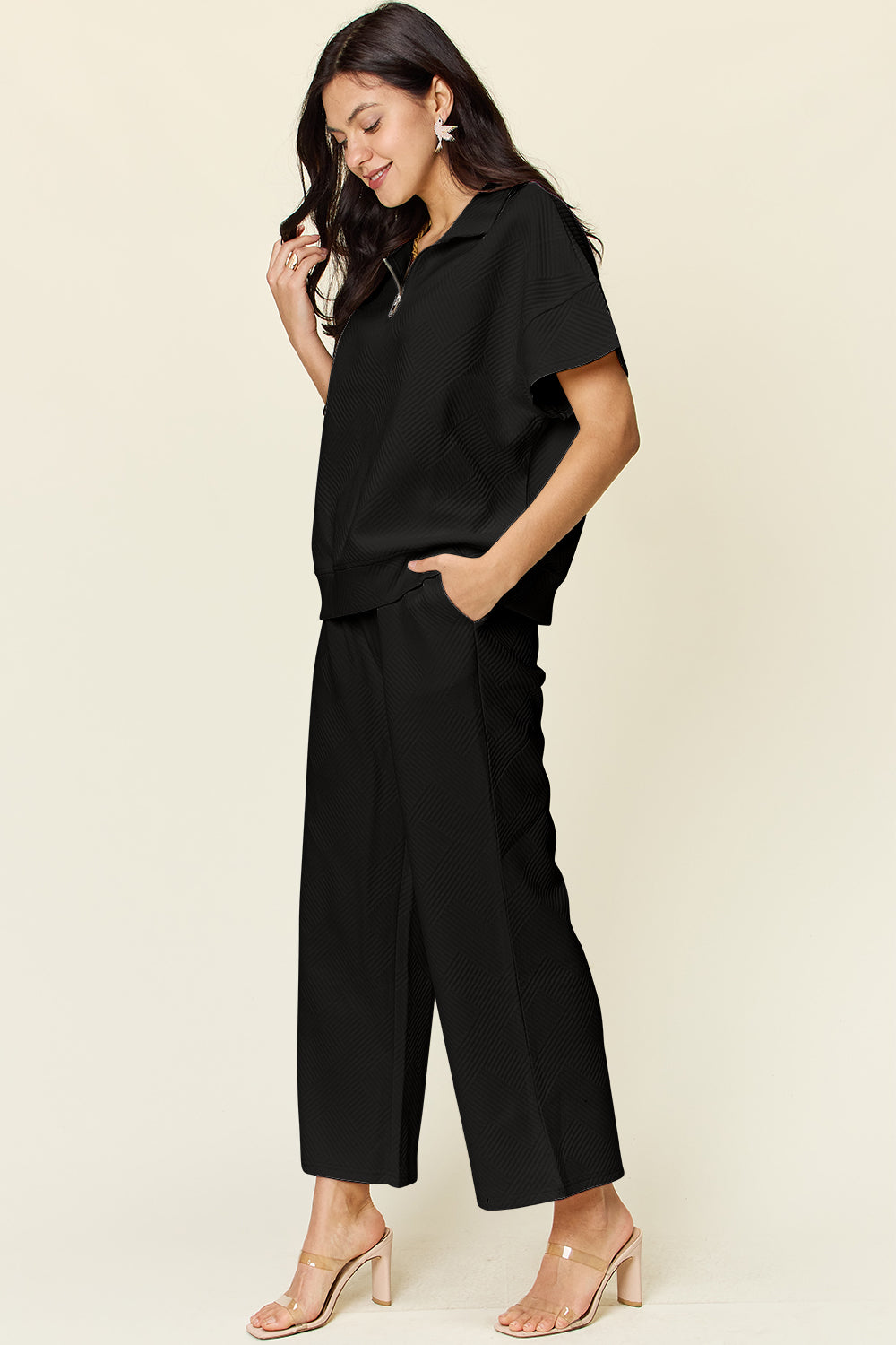 A woman is wearing the Double Take Full Size Texture Half Zip Short Sleeve Top and Pants Set, featuring a white zip-up short-sleeve top and matching wide-leg, drawstring pants. She stands with one hand in her pocket and smiles against a light background.