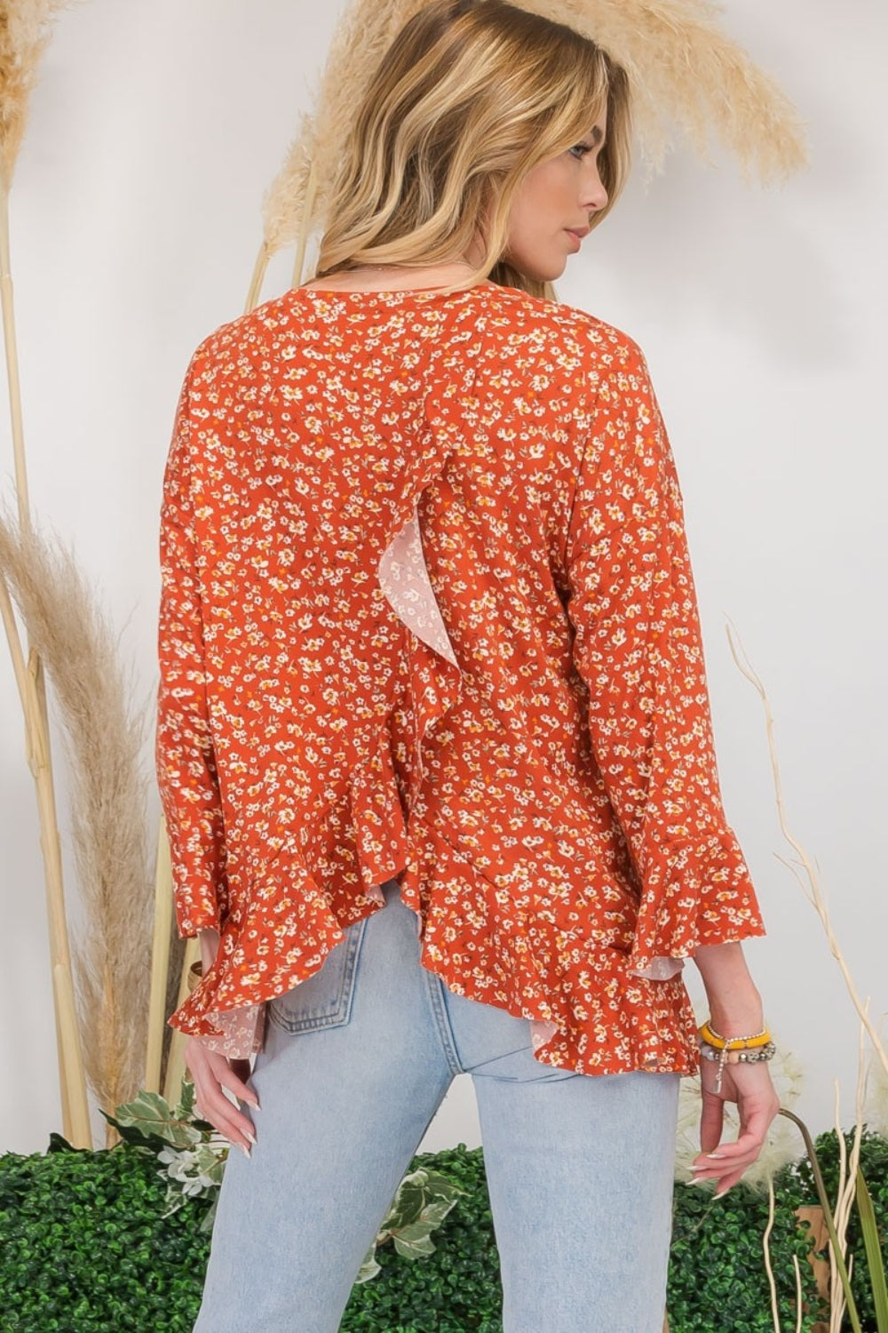 A woman wearing the Celeste Full Size Floral Ruffle Detail Top and jeans stands smiling in front of pampas grass and greenery, exuding feminine flair.