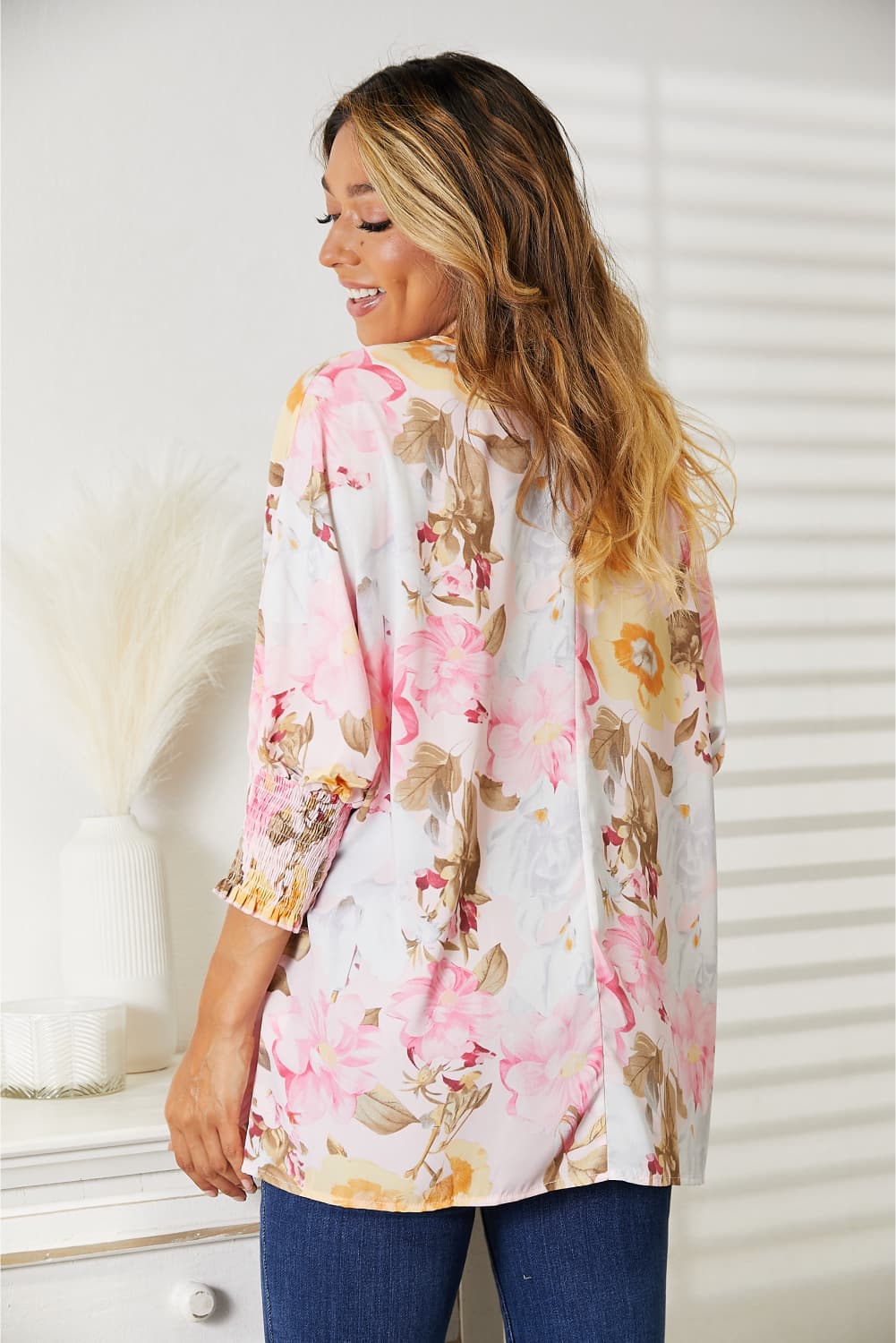 A woman in a Double Take Floral Round Neck Three-Quarter Sleeve Top and jeans stands smiling in a brightly lit room with a white background and drawers on the side.