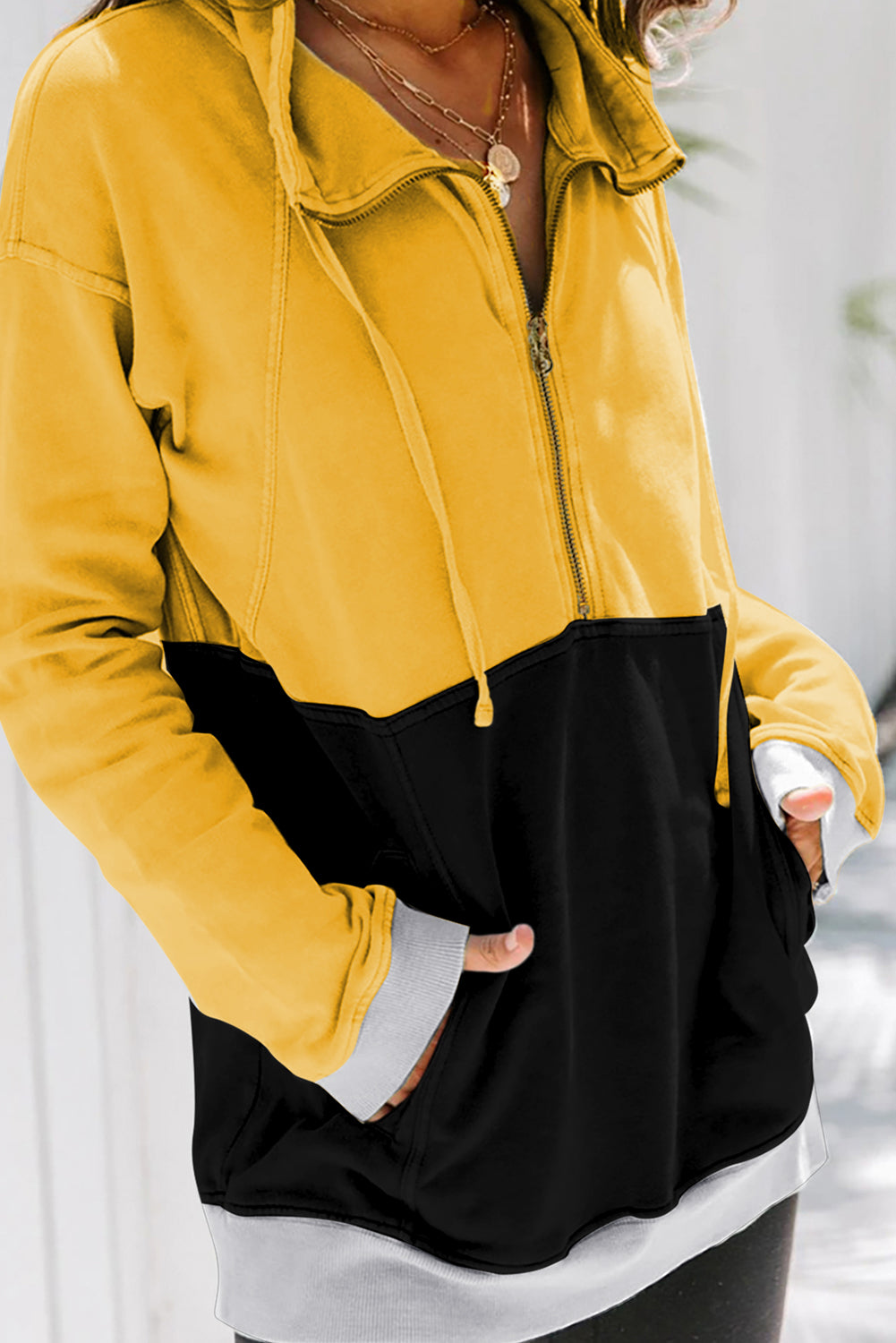 A person with long, light hair in a ponytail, wearing a Black Zipped Colorblock Sweatshirt with Pockets, blue jeans, and glasses, is holding a white cup and a brown handbag, shown from behind against a gray wall.